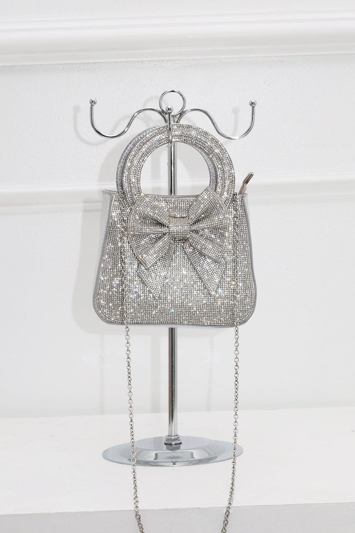 EDGE — silver rhinestone bow-embellished mini clutch with chain strap, styled on a display stand