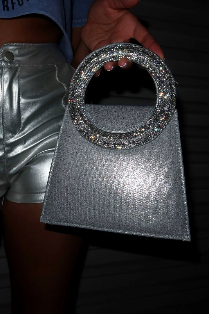 EDGE — silver satin top handle evening bag with a rhinestone circular handle, held by a model